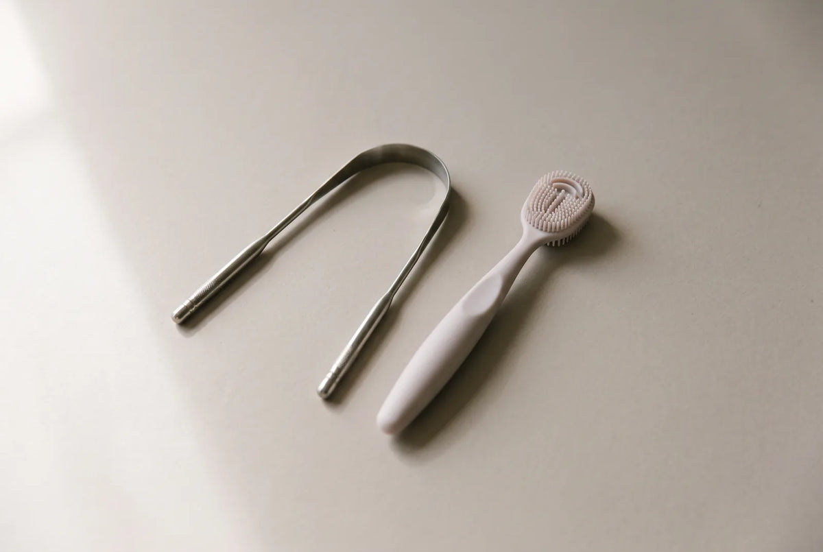 Racleur à langue en inox et brosse à dents à poils souples posés côte à côte sur le plan d'une salle de bain