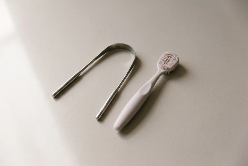 Racleur à langue en inox et brosse à dents à poils souples posés côte à côte sur le plan d'une salle de bain